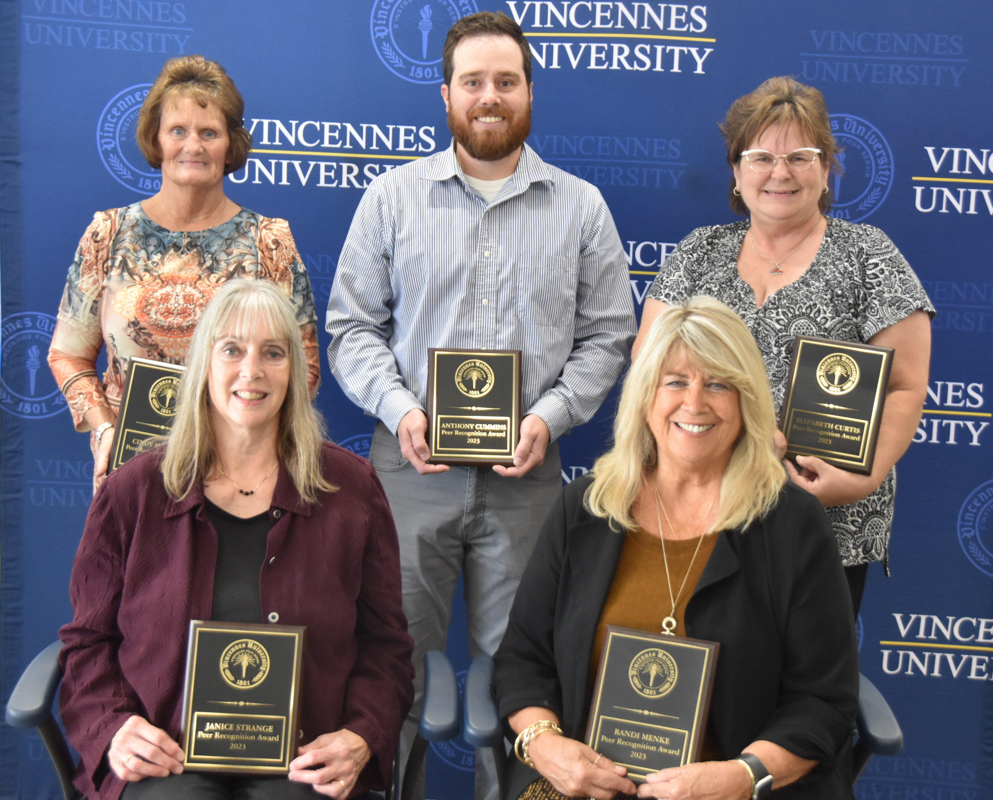 Four female and one male VU staff and faculty holding their Peer Recognition Awards standing and sitting in front of a VU step and repeat backdrop with a VU logo.